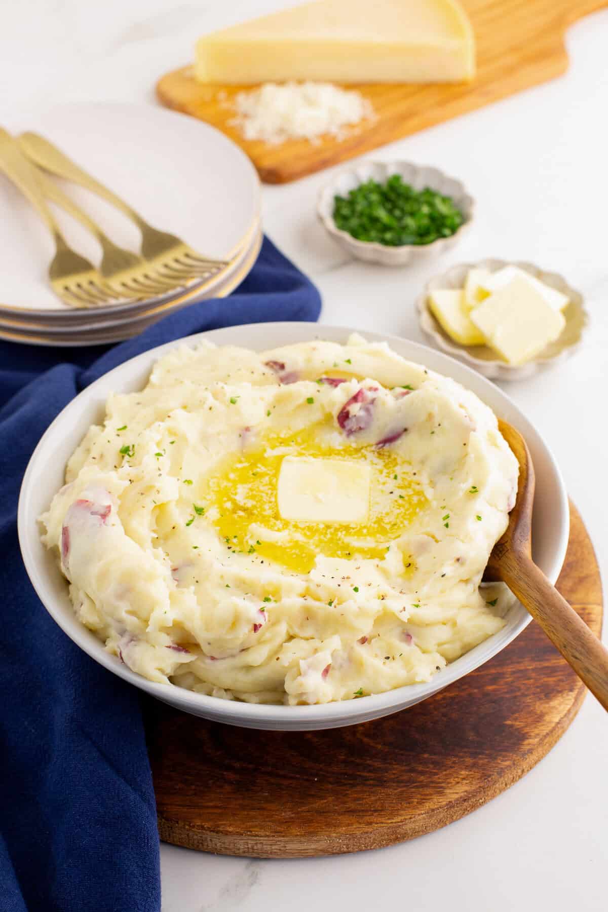 White bowl filled with red skin mashed potatoes, topped with a pat of melting butter and sprinkled with herbs, set on a wooden board with plates, forks, Parmesan cheese wedge, chopped chives, and extra butter in the background.