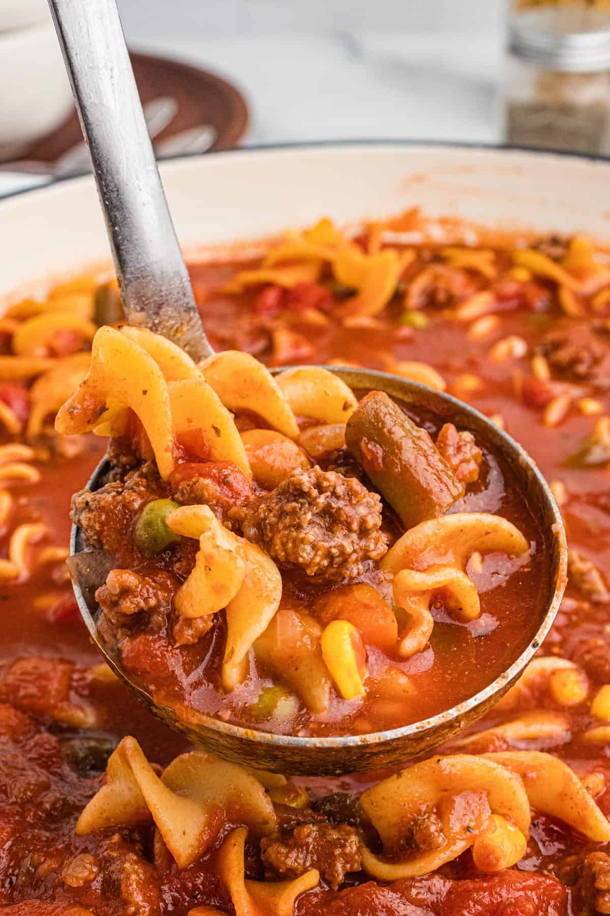 Close-up of a ladle full of hamburger noodle soup showing egg noodles, ground beef chunks, corn, carrots, green beans, peas, and tomatoes in a red broth.