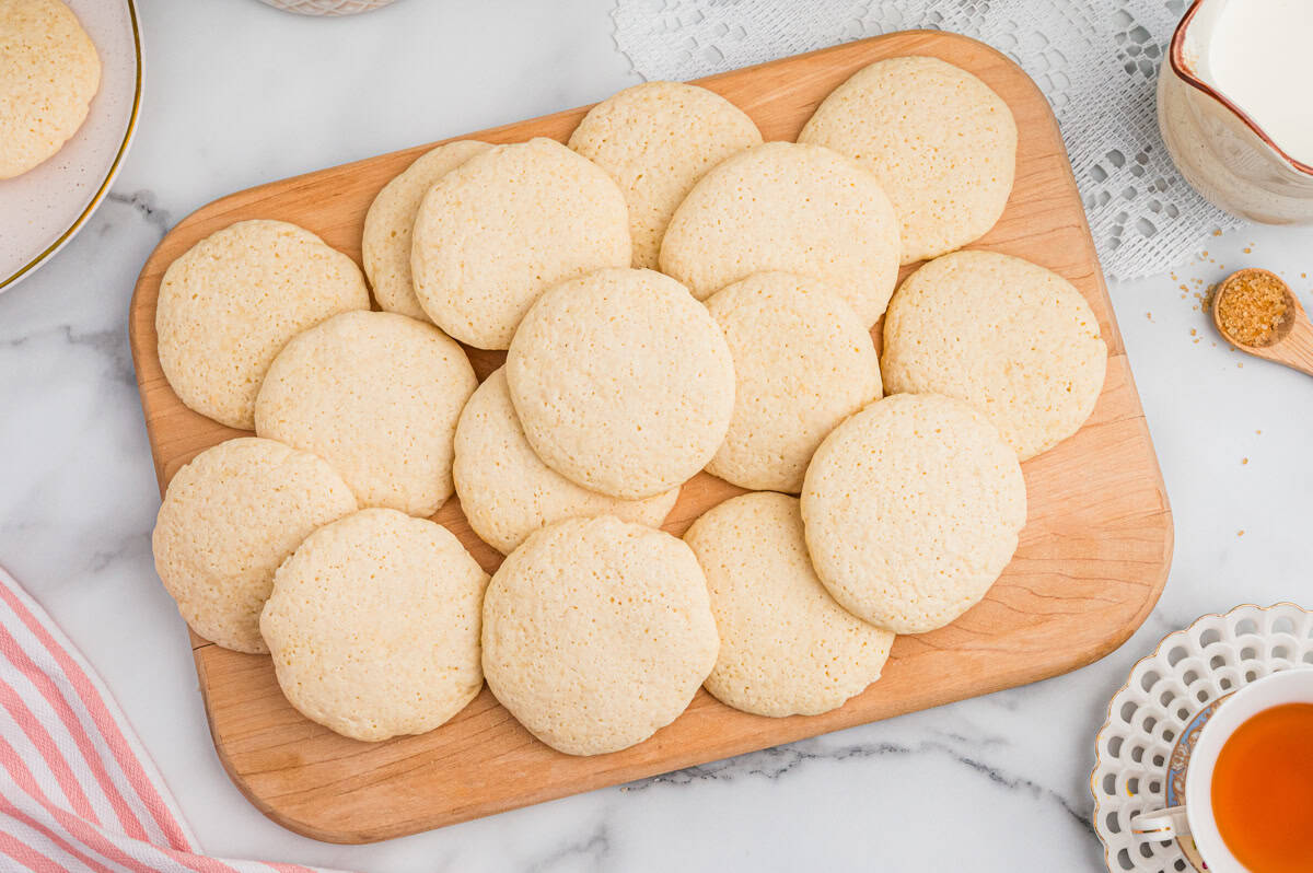Overhead view of a batch of old-fashioned tea cakes arranged on a rectangular wooden board. The cookies are pale golden and round, placed on a marble surface with lace and ceramic dishes nearby.