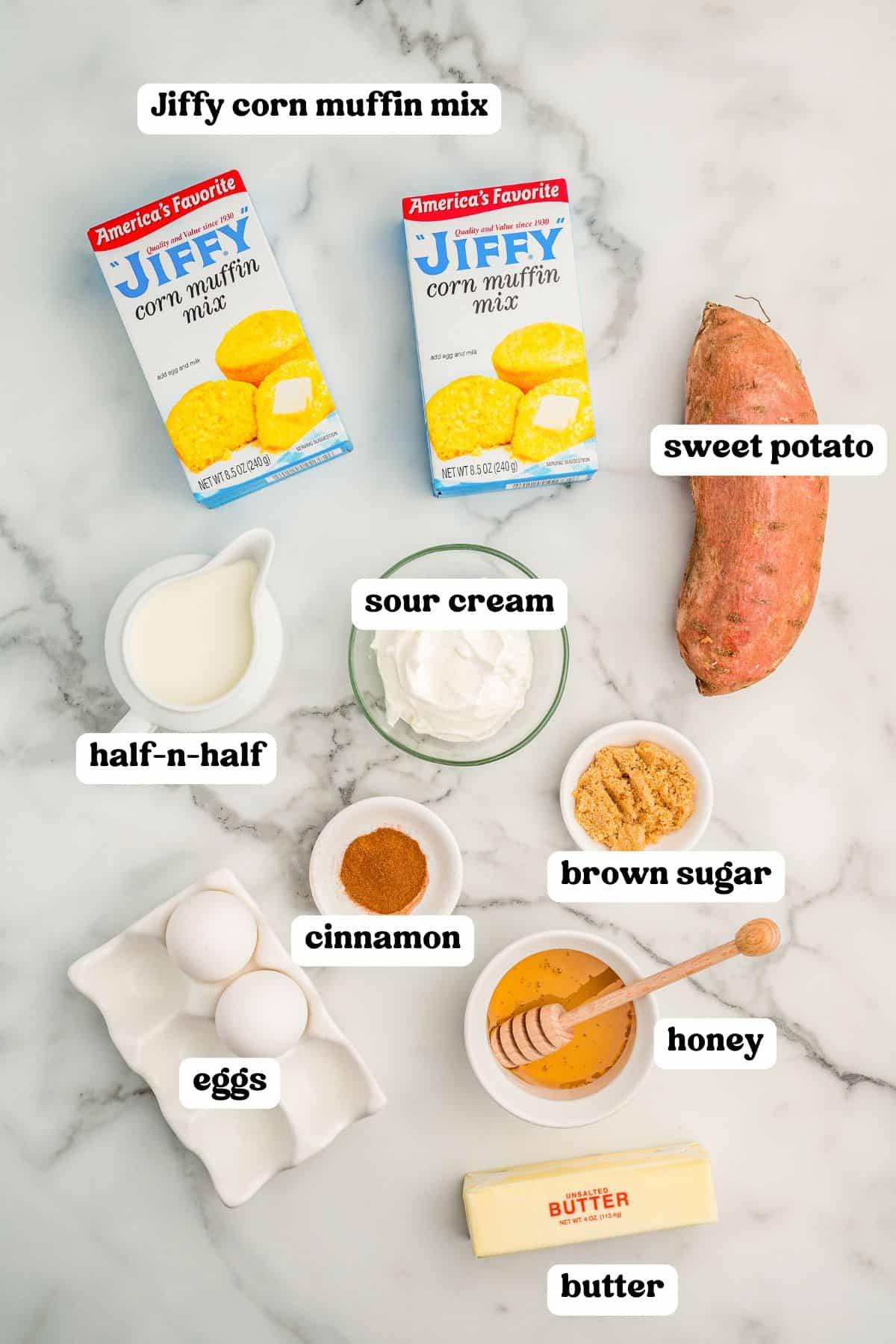 Labeled ingredients for sweet potato cornbread including two boxes of Jiffy corn muffin mix, a sweet potato, sour cream, half-and-half, cinnamon, brown sugar, honey with a dipper, butter, and two eggs.
