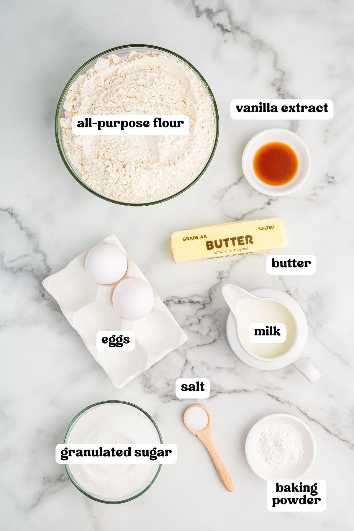 Labeled ingredient for making old-fashioned tea cakes. Items include a bowl of all-purpose flour, two eggs in a ceramic holder, granulated sugar, a stick of salted butter, a small bowl of vanilla extract, milk in a white pitcher, salt in a wooden spoon, and baking powder in a small bowl.