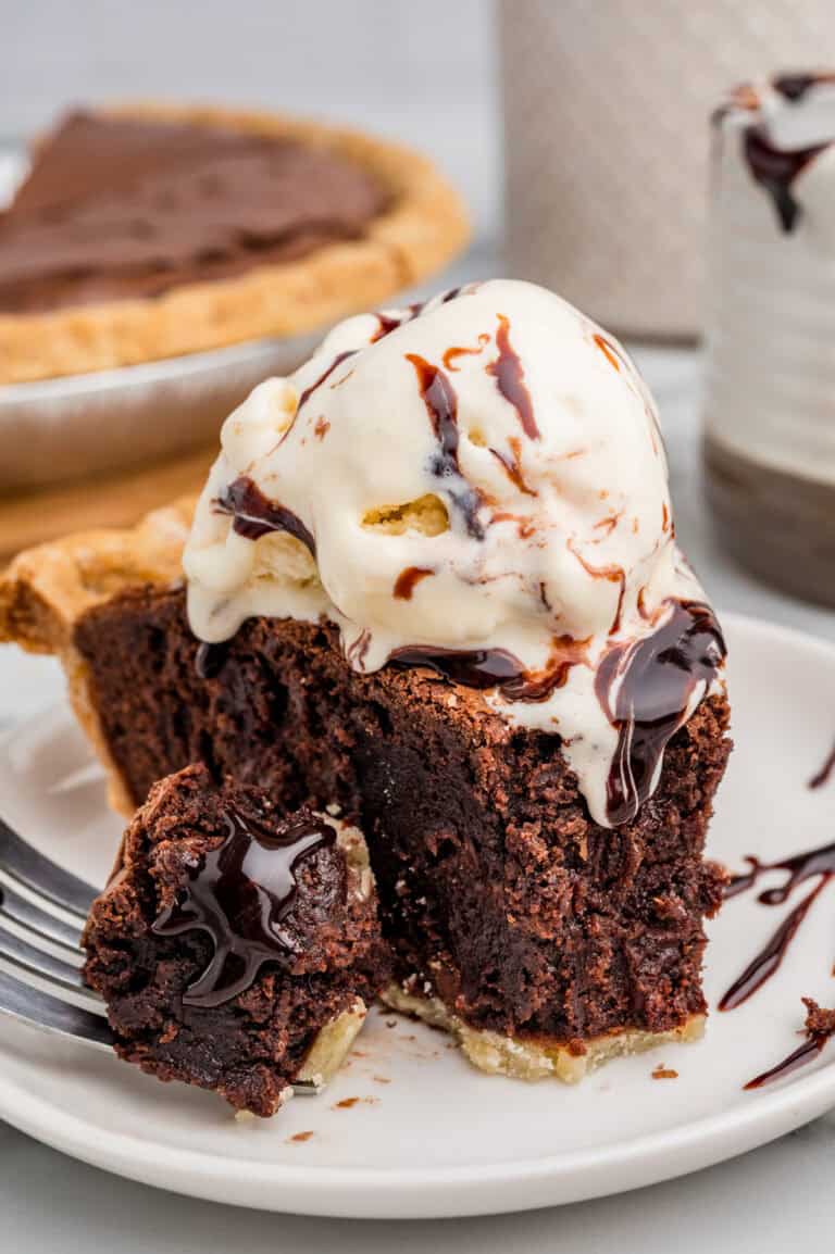 Angle view of a brownie pie slice with ice cream and chocolate syrup. A bite sits on a fork in front of the slice, showing gooey melted chocolate inside.