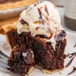 Slice of chocolate brownie pie with ice cream and chocolate syrup. A bite sits on a fork in front of the slice, showing gooey melted chocolate inside.
