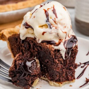 Slice of chocolate brownie pie with ice cream and chocolate syrup. A bite sits on a fork in front of the slice, showing gooey melted chocolate inside.