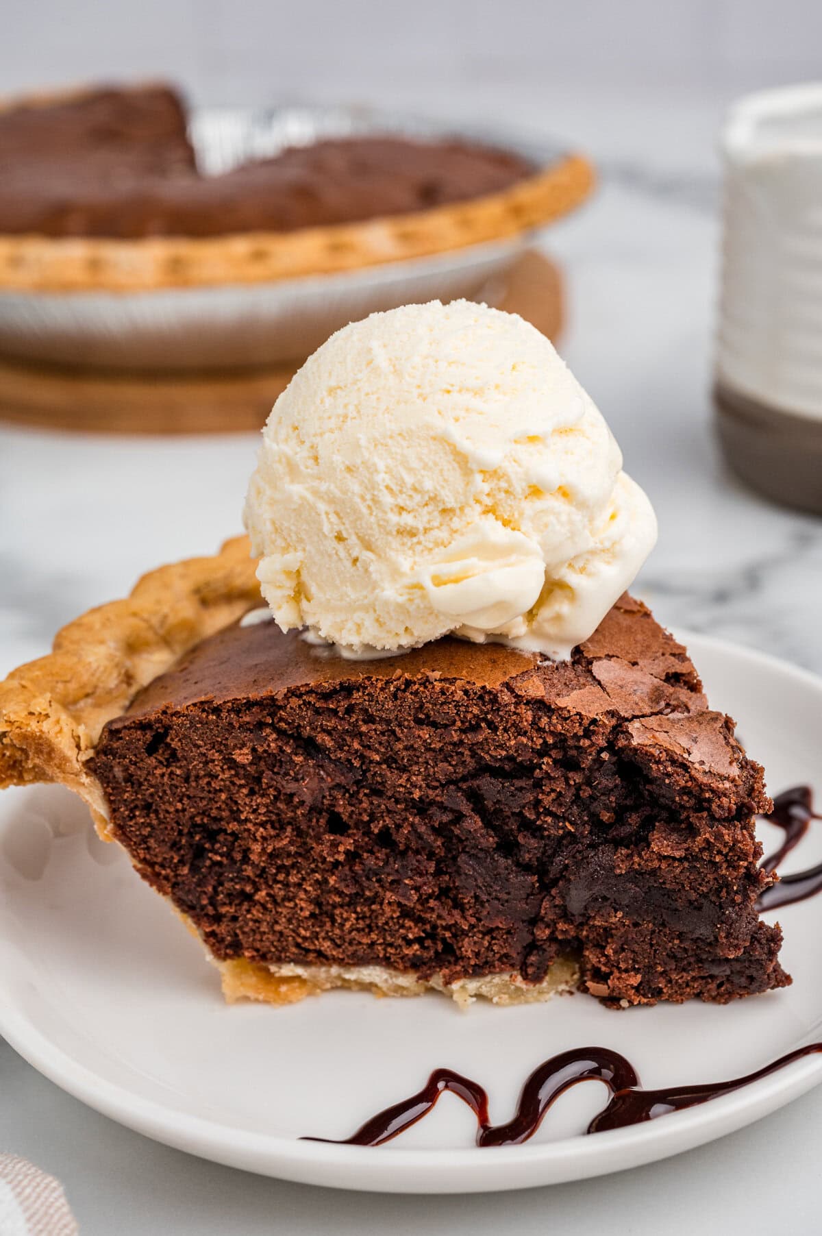 A slice of brownie pie served on a white plate, topped with a scoop of vanilla ice cream. Chocolate syrup is drizzled on the plate, and the rest of the pie is in the background.