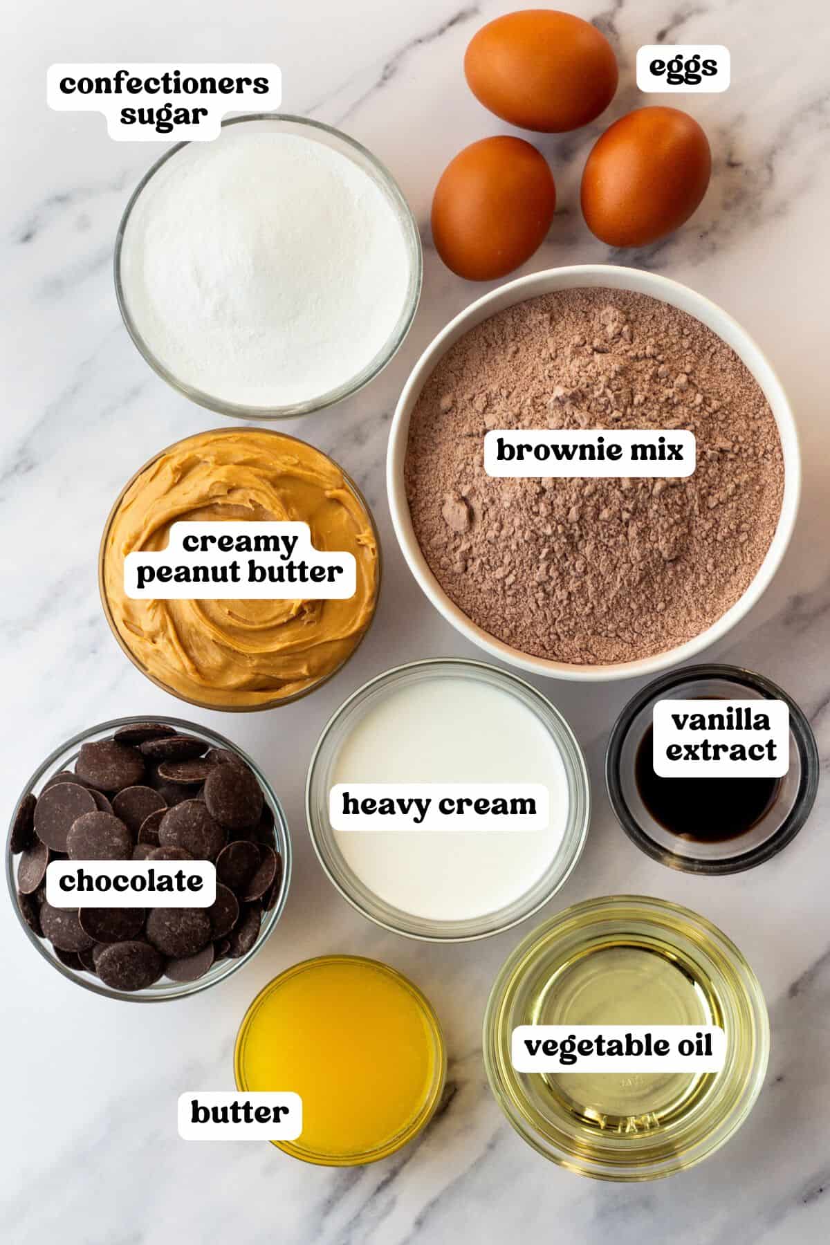 Flat lay of labeled ingredients in bowls for buckeye brownies, arranged on a marble surface. Items include: confectioners sugar, creamy peanut butter, chocolate, heavy cream, butter, vegetable oil, vanilla extract, brownie mix, and three eggs.