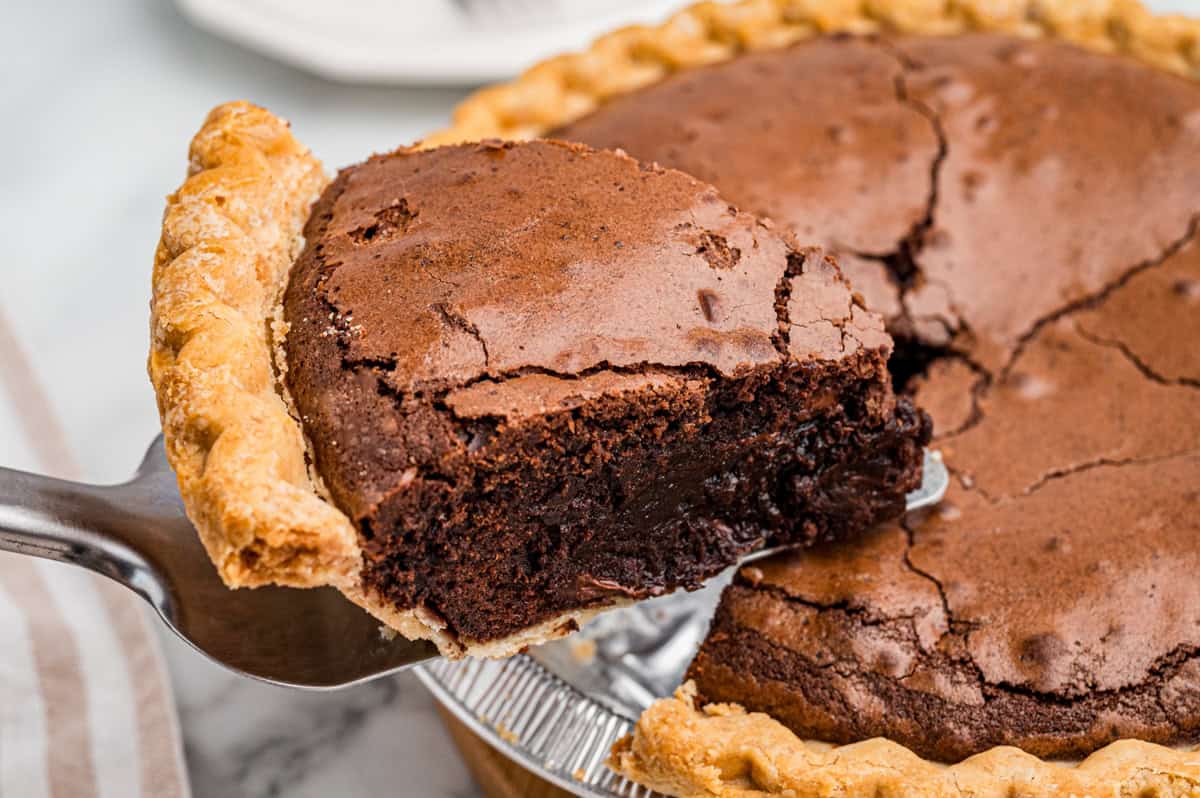 Close-up of a slice of brownie pie being lifted from the pie dish. The pie has a cracked top and rich, fudgy center with a golden crust.