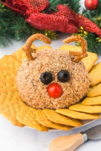 A festive reindeer cheese ball decorated with pretzel antlers, black olive eyes, and a red cherry tomato nose. The cheese ball is coated in chopped nuts and surrounded by golden crackers on a white plate with holiday decorations in the background.