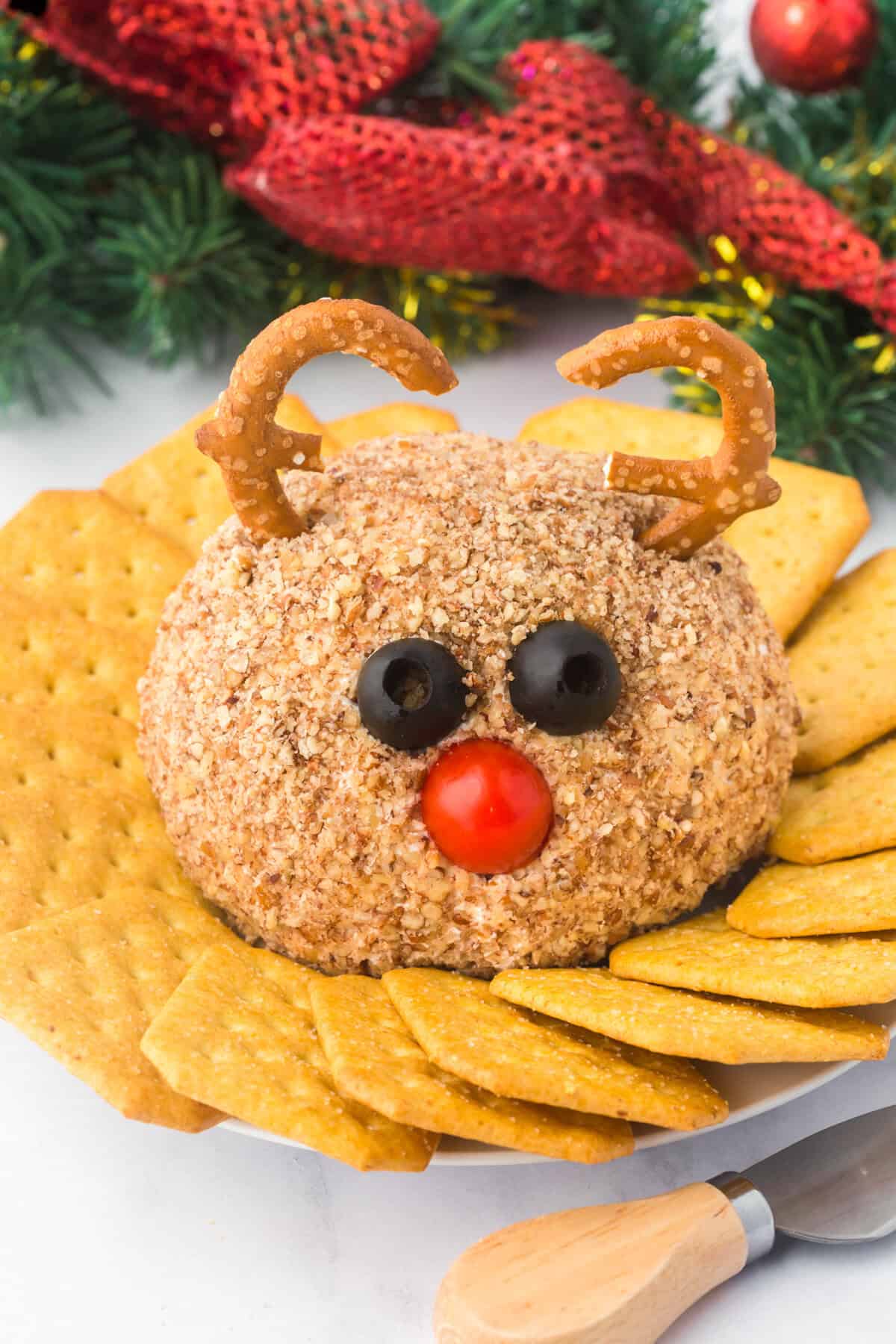 A festive reindeer cheese ball decorated with pretzel antlers, black olive eyes, and a red cherry tomato nose. The cheese ball is coated in chopped nuts and surrounded by golden crackers on a white plate with holiday decorations in the background.