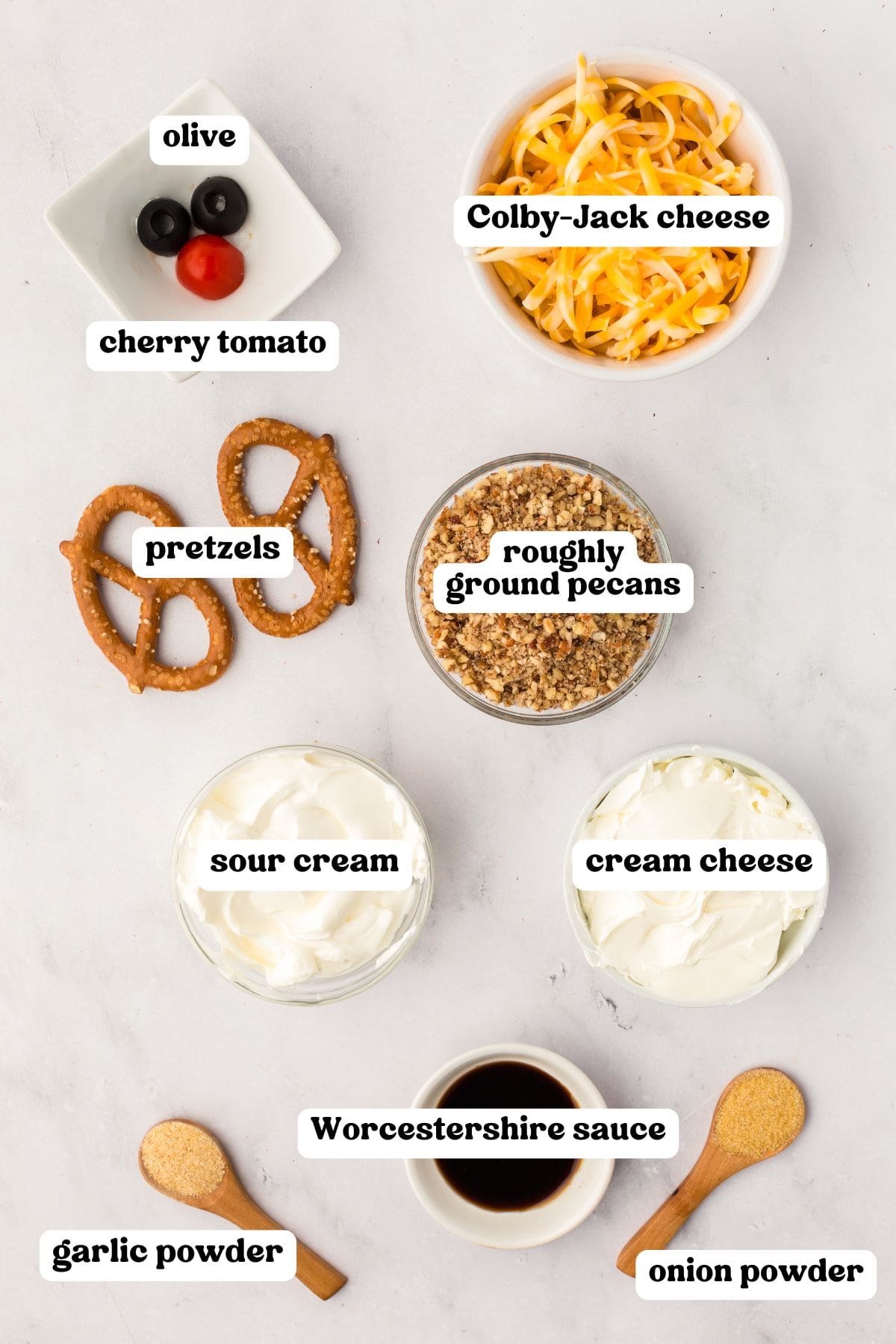 Overhead view of labeled ingredients for a reindeer cheese ball including black olives, a cherry tomato, shredded Colby-Jack cheese, two pretzels, roughly ground pecans, sour cream, cream cheese, Worcestershire sauce, garlic powder, and onion powder.