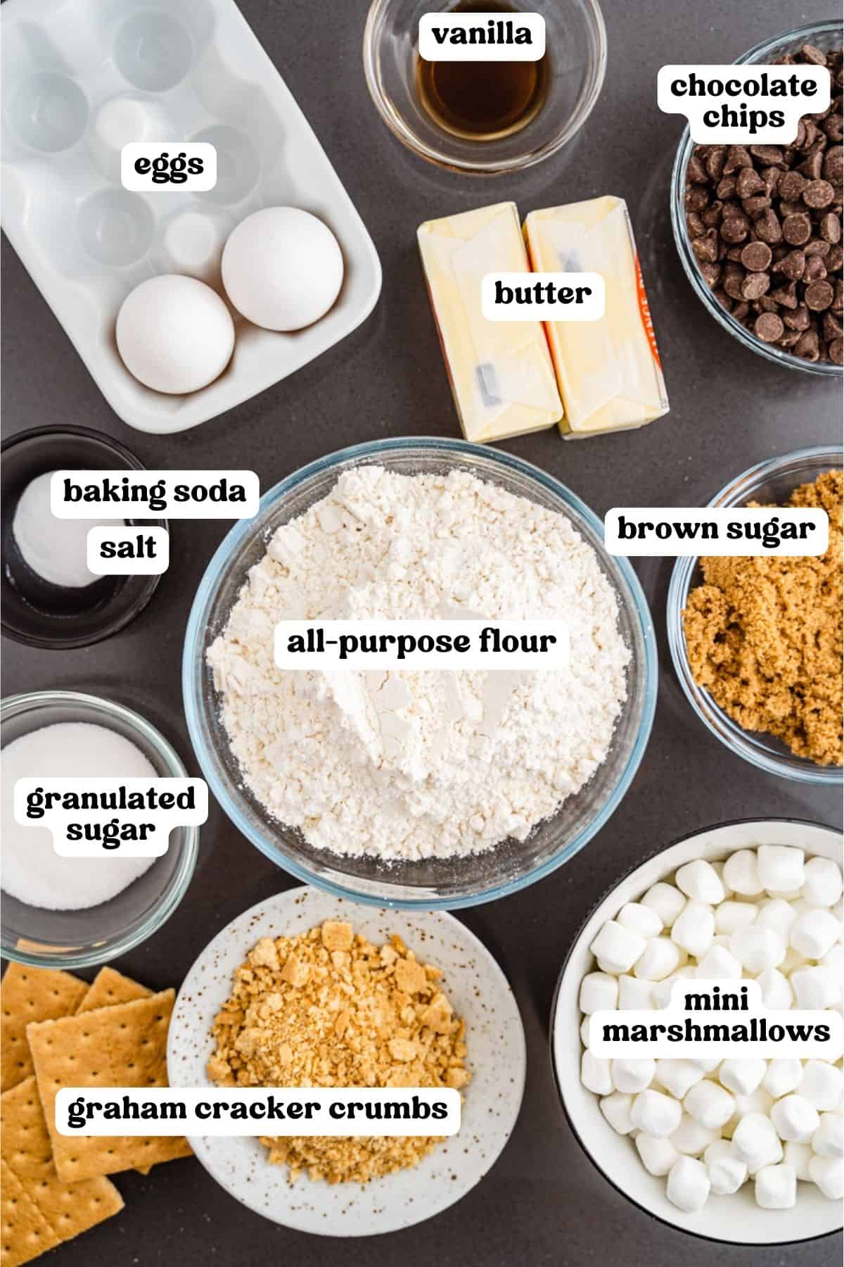 Labeled baking ingredients in bowls for s’mores cookies, including flour, brown sugar, chocolate chips, butter, eggs, vanilla, graham cracker crumbs, mini marshmallows, granulated sugar, salt, and baking soda.
