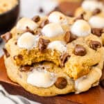 Two stacked s'mores cookies on a wooden board, with the top cookie missing a bite, showing a soft, golden interior and melted marshmallows and chocolate chips on top.