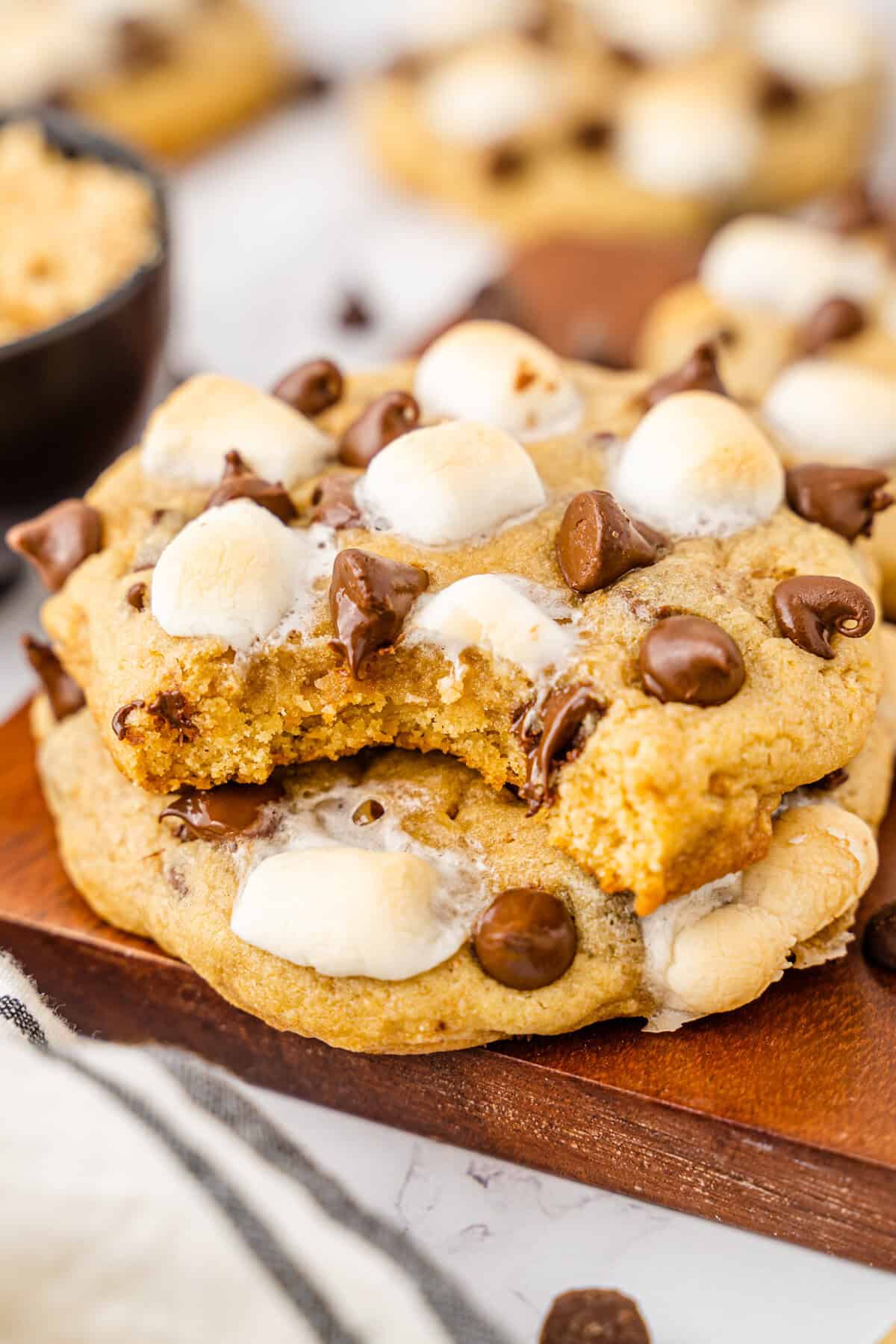 Two stacked s'mores cookies on a wooden board, with the top cookie missing a bite, showing a soft, golden interior and melted marshmallows and chocolate chips on top.