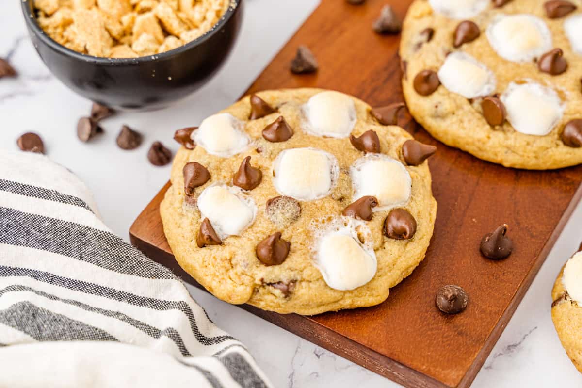 S'mores chocolate chip cookies on wooden board with chocolate chips scattered around them.