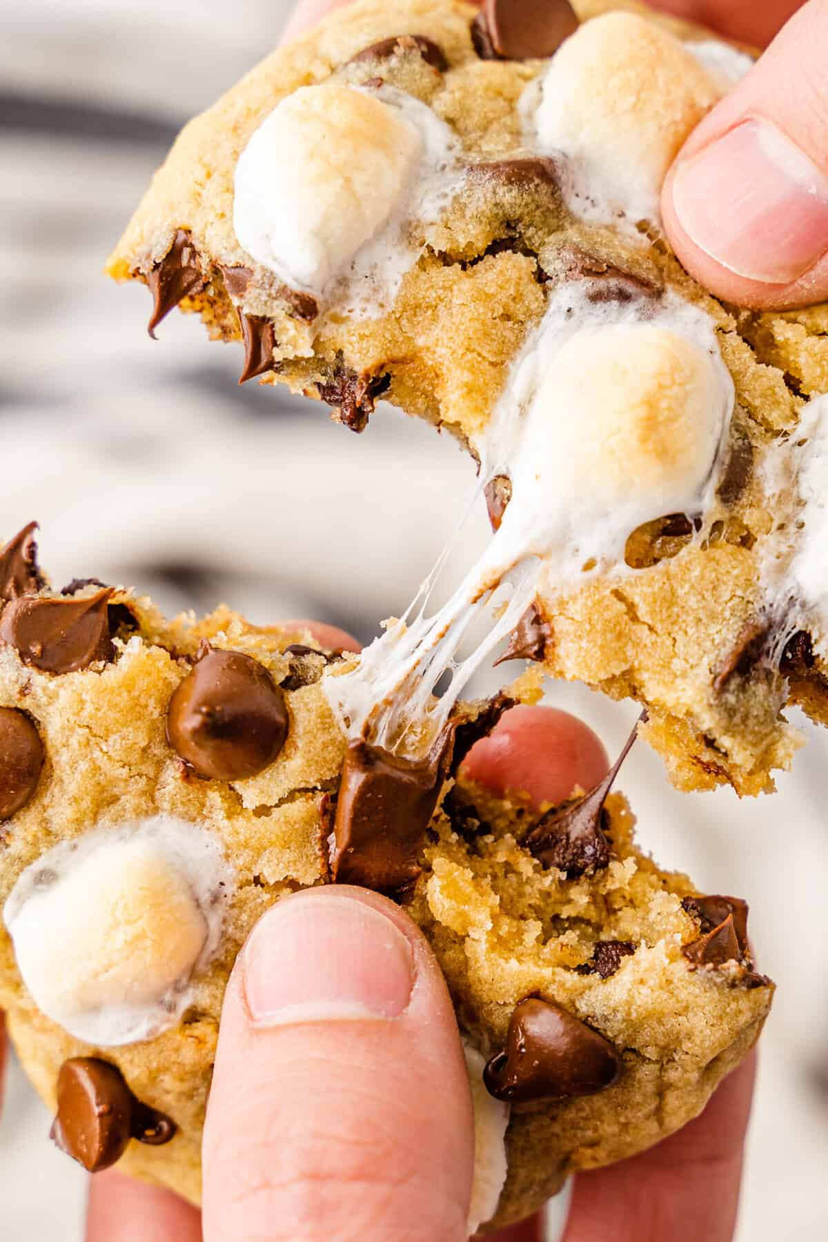 Fingers pulling apart a s'mores chocolate chip cookie, showing melted chocolate chips and gooey marshmallow strands stretching between halves.