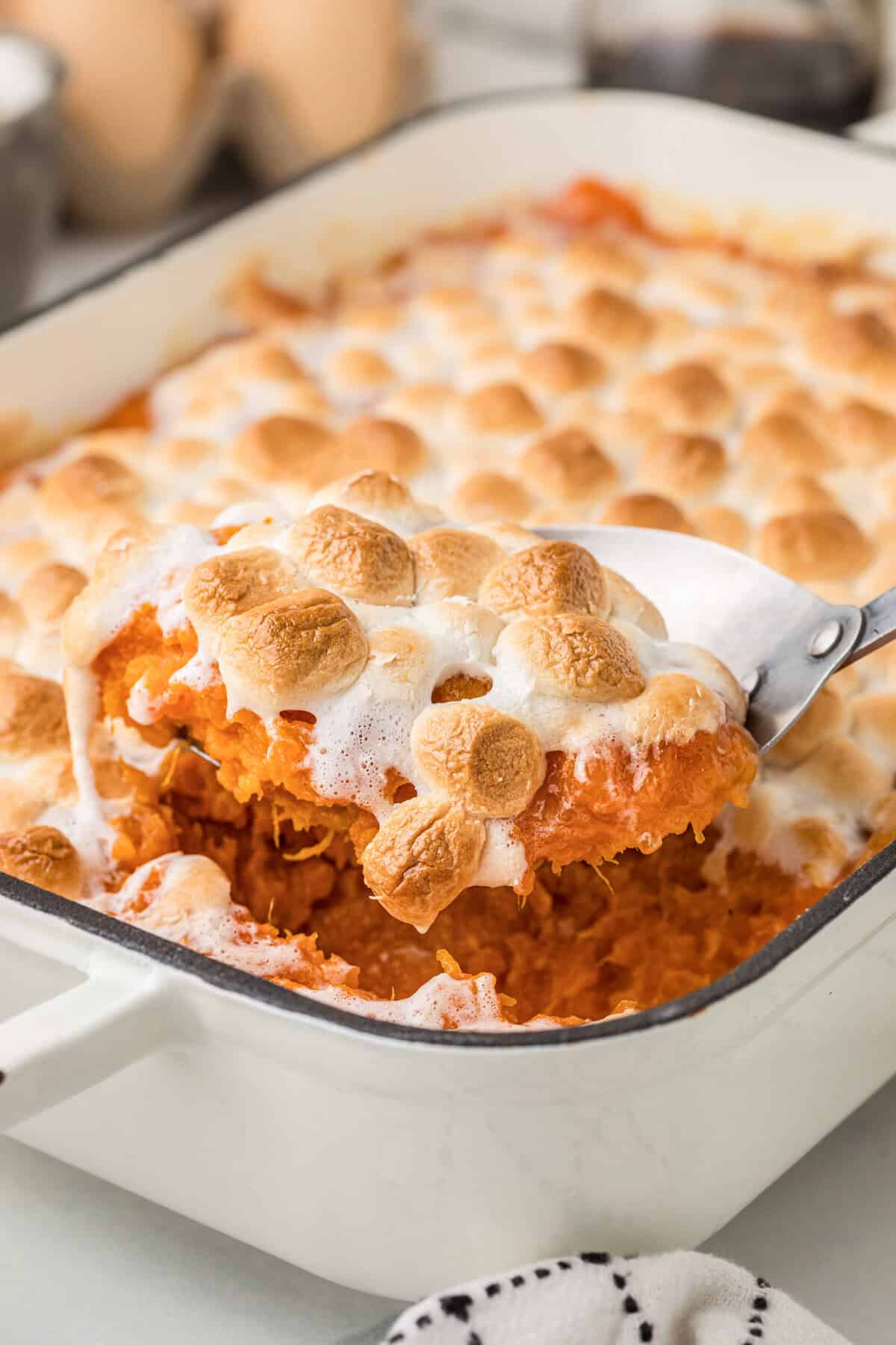 A spoon lifting a portion of sweet potato casserole with marshmallows from a white baking dish, showing fluffy orange sweet potatoes topped with golden-brown toasted mini marshmallows.