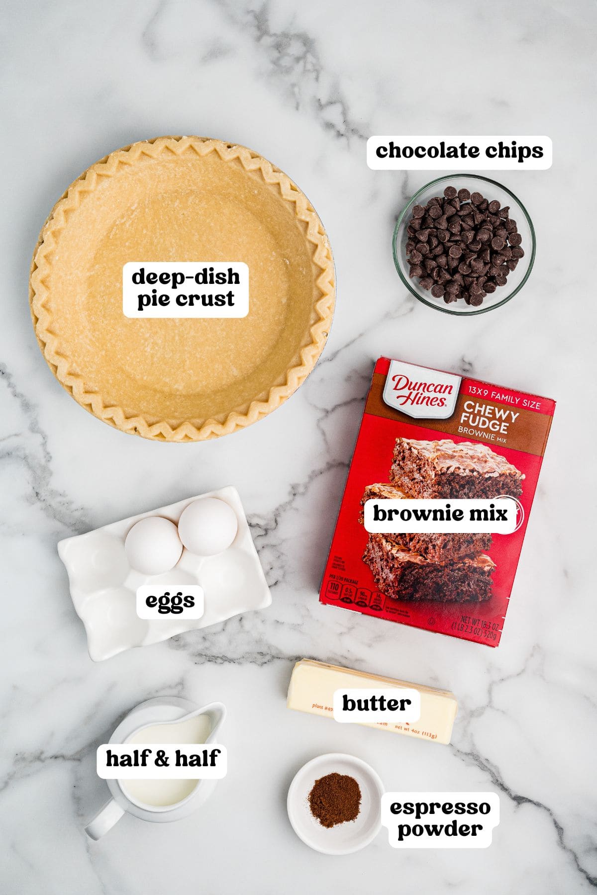 Flat lay of ingredients for brownie pie, each labeled with text. Items include a deep-dish pie crust, chocolate chips, a box of Duncan Hines Chewy Fudge Brownie Mix, two eggs, a stick of butter, half & half, and espresso powder.