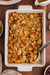 A 9x13 baking dish filled with homemade bread stuffing. Surrounding it are bowls of green beans and cranberry sauce, a gravy boat, salt and pepper shakers, and part of a roasted turkey on a serving platter.