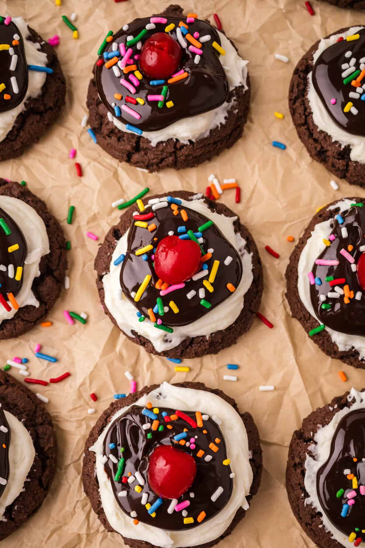 Top-down view of Brownie Sundae cookies arranged on parchment paper, each topped with white frosting, a layer of chocolate sauce, rainbow sprinkles, and a red cherry, with extra sprinkles scattered around.