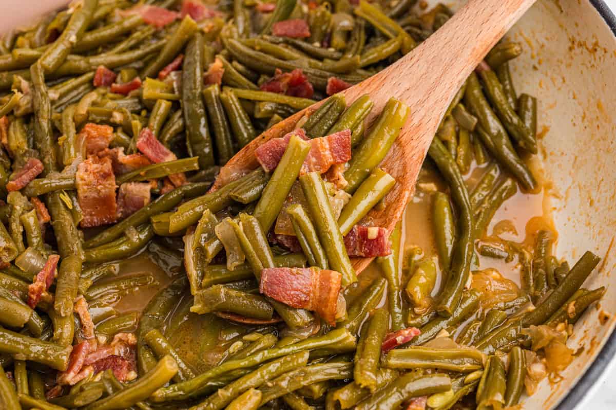 Close-up of green beans and bacon in a shallow pan with broth. A wooden spoon lifts a portion, showing chopped bacon, onions, and soft green beans coated in sauce.