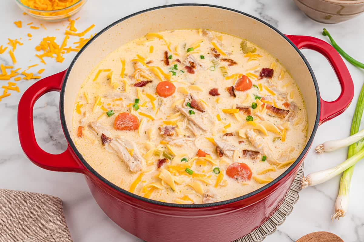 A red Dutch oven filled with creamy chicken noodle soup, featuring egg noodles, shredded chicken, bacon pieces, carrot slices, green onions, and melted cheese.
