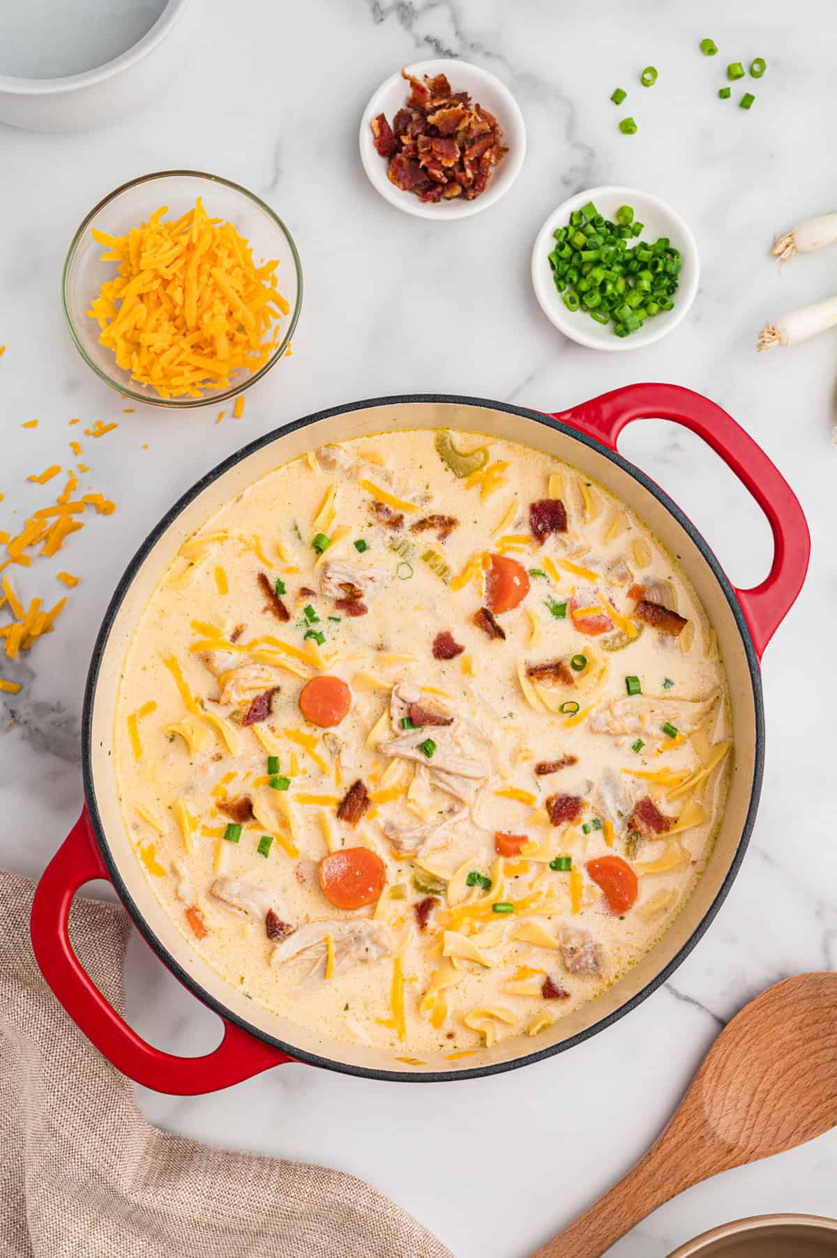 Overhead image of crack chicken noodle soup in a Dutch oven. Surrounding the pot are bowls of shredded cheddar, crumbled bacon, and chopped green onions, and whole green onions.