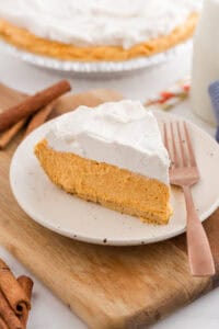 slice of no-bake pumpkin pie with a graham cracker crust and whipped topping is placed on a white plate with a bronze fork.