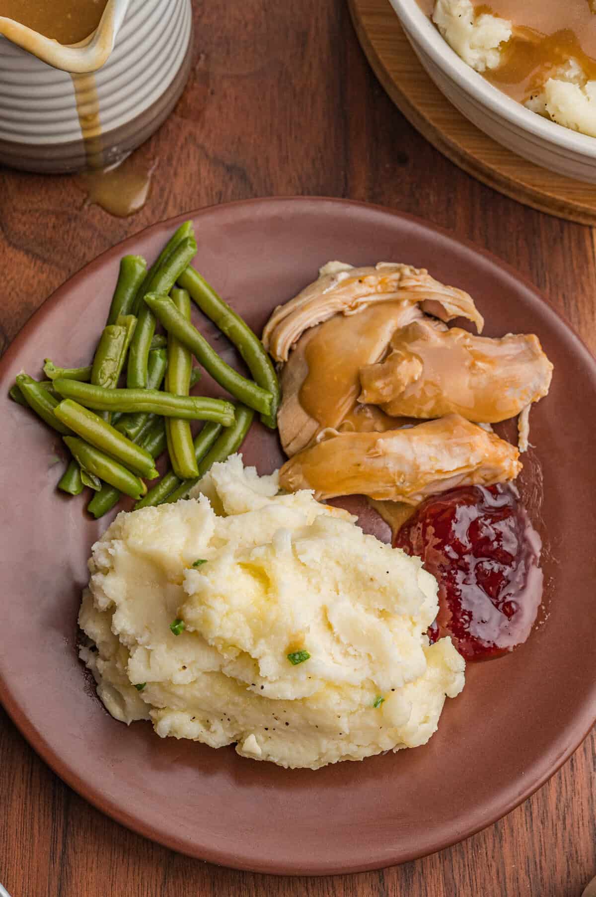 Dinner plate containing mashed potatoes with chives and melted butter, cooked green beans, turkey slices with brown gravy, and cranberry sauce.