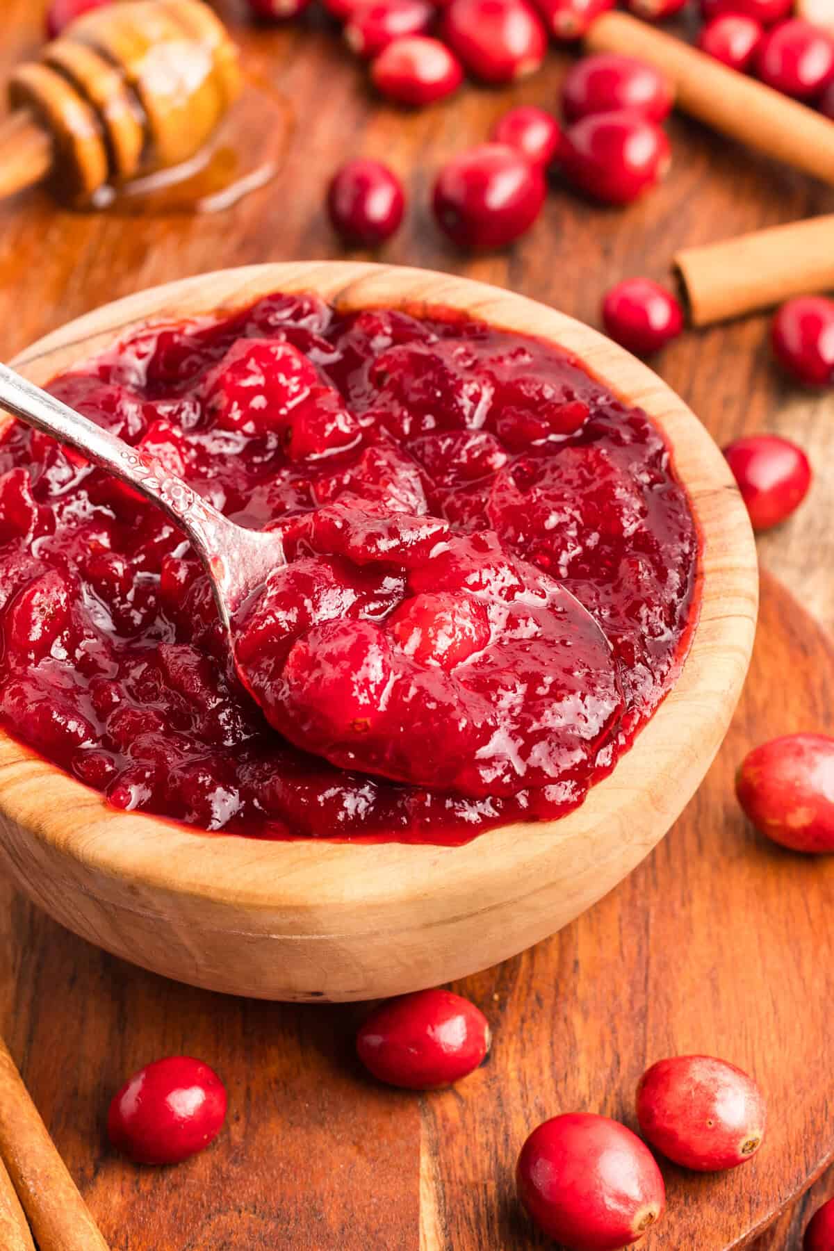A close-up of a silver spoon lifting a scoop of thick, chunky cranberry sauce from a wooden bowl. The sauce is glossy and vibrant red.
