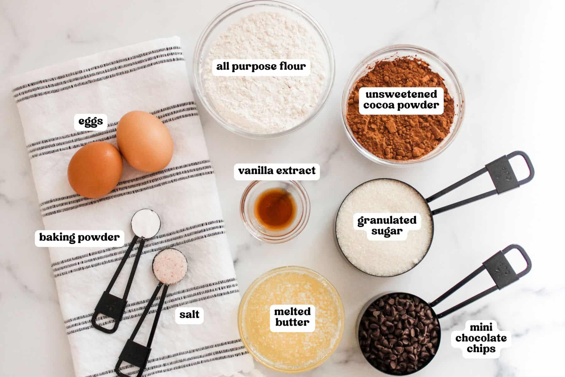 Flat lay of brownie bite ingredients on a white surface with black measuring spoons and a striped towel. Ingredients are labeled: two eggs, baking powder, salt, all-purpose flour, unsweetened cocoa powder, vanilla extract, granulated sugar, melted butter, and mini chocolate chips.