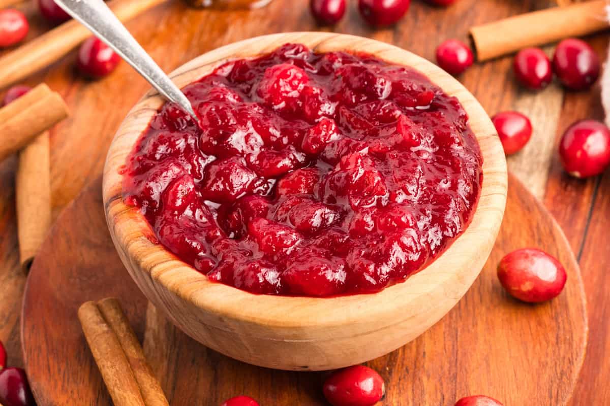 Spoon resting in a bowl of thick cranberry sauce with whole cranberries. Cinnamon sticks and fresh cranberries are spread about the bowl.