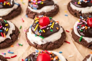 Hot fudge sundae cookies made with a chocolate brownie cookie base topped with white frosting, chocolate sauce, rainbow sprinkles, and a cherry.