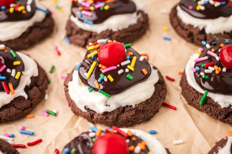 Hot fudge sundae cookies made with a chocolate brownie cookie base topped with white frosting, chocolate sauce, rainbow sprinkles, and a cherry.