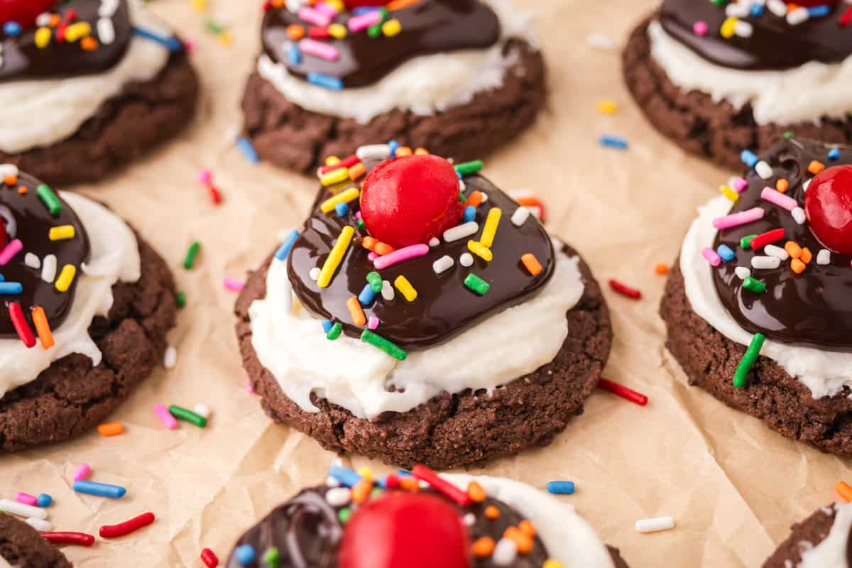 Hot fudge sundae cookies made with a chocolate brownie cookie base topped with white frosting, chocolate sauce, rainbow sprinkles, and a cherry.
