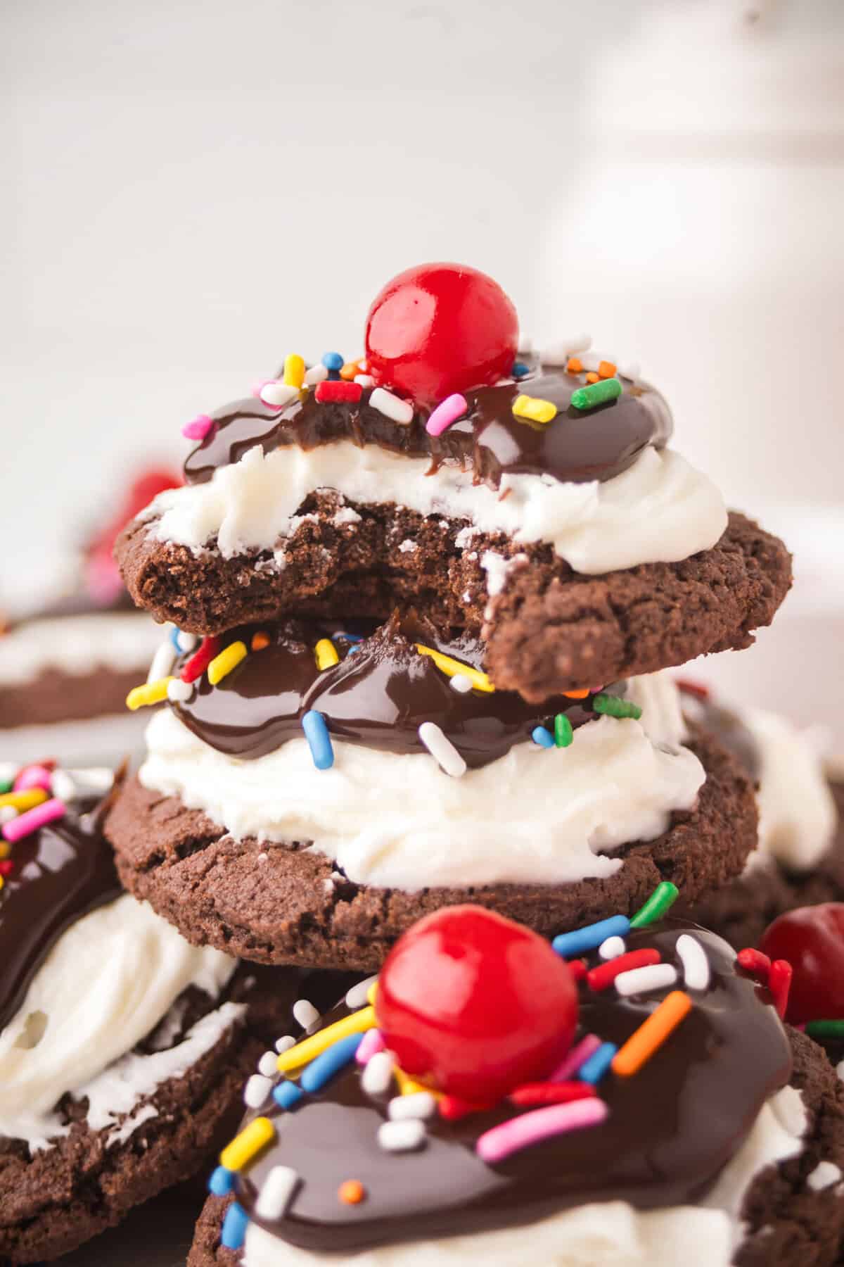 Ice cream sundae brownie cookies stacked with frosting, chocolate, sprinkles, and cherries; top cookie has a bite taken out.