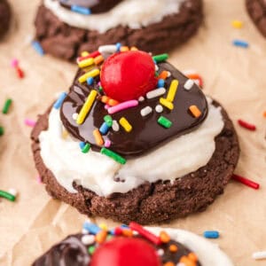 Hot fudge sundae cookie with a chocolate cookie base topped with white frosting, hot fudge sauce, rainbow sprinkles, and a maraschino cherry.