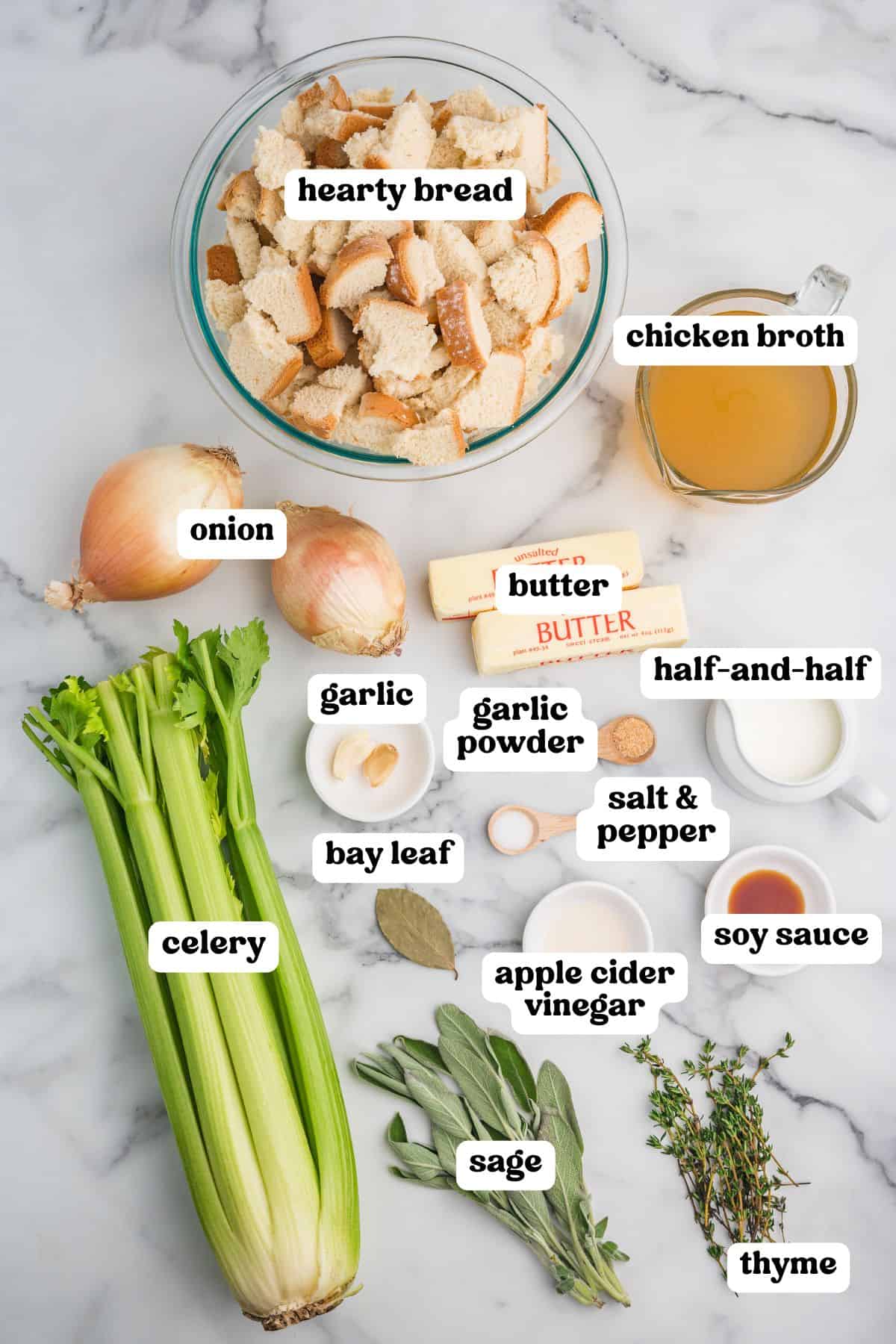Homemade stuffing ingredients labeled on a marble surface. Visible items include a bowl of torn hearty bread, chicken broth, onions, celery, garlic, bay leaf, butter, half-and-half, garlic powder, salt and pepper, soy sauce, apple cider vinegar, fresh sage, and thyme.