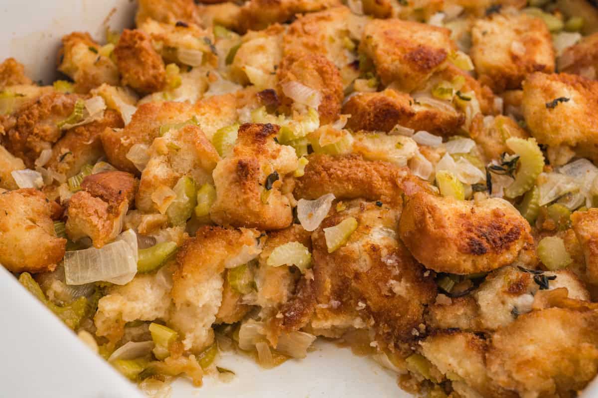 A detailed close-up of old-fashioned homemade stuffing in a white dish, showing crispy golden-brown bread cubes mixed with chopped celery, onions, and herbs.
