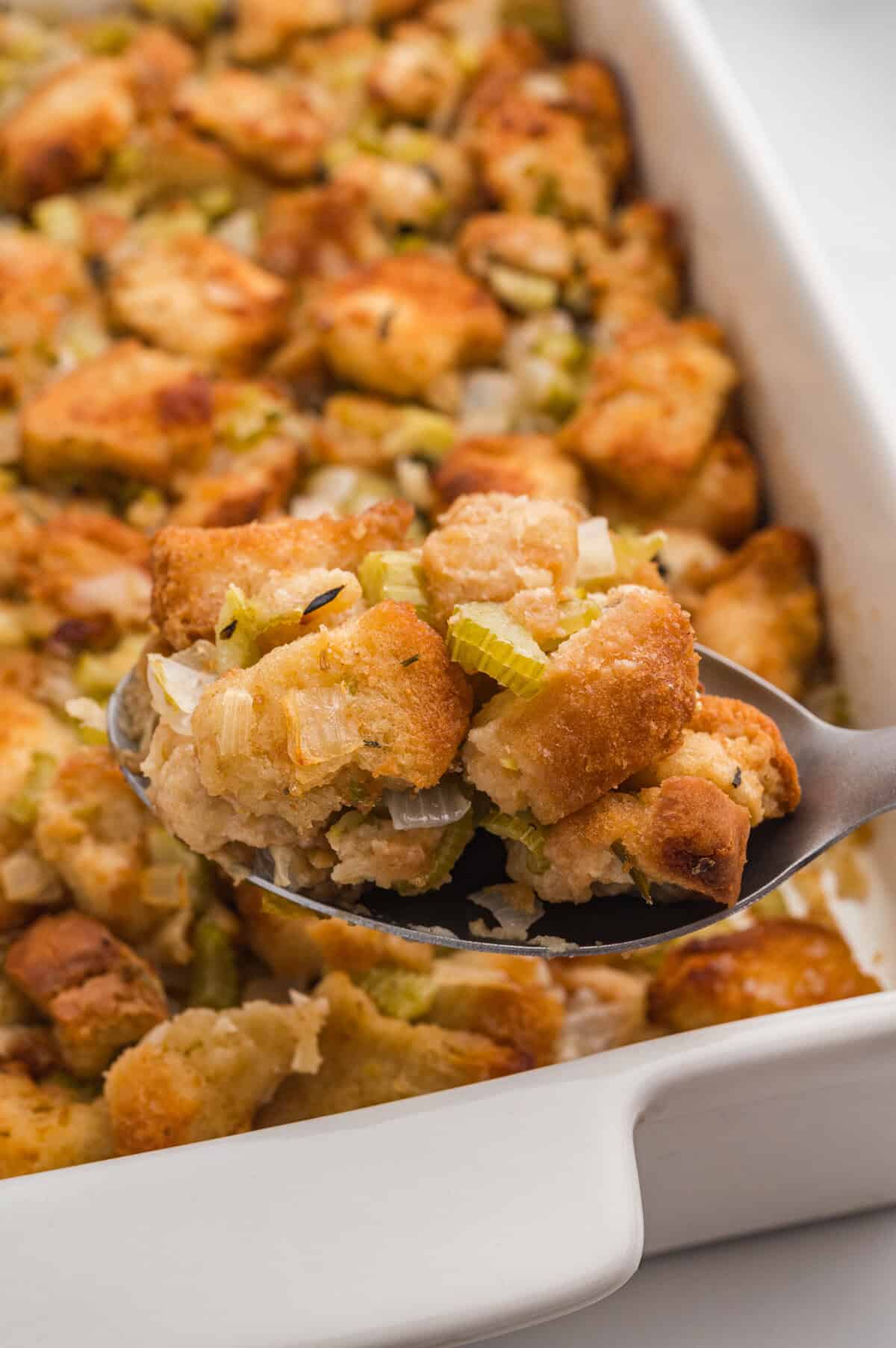 A close-up of a spoonful of homemade stuffing lifted above a white baking dish. The spoon holds rustic toasted bread cubes mixed with chopped celery, onions, and herbs.