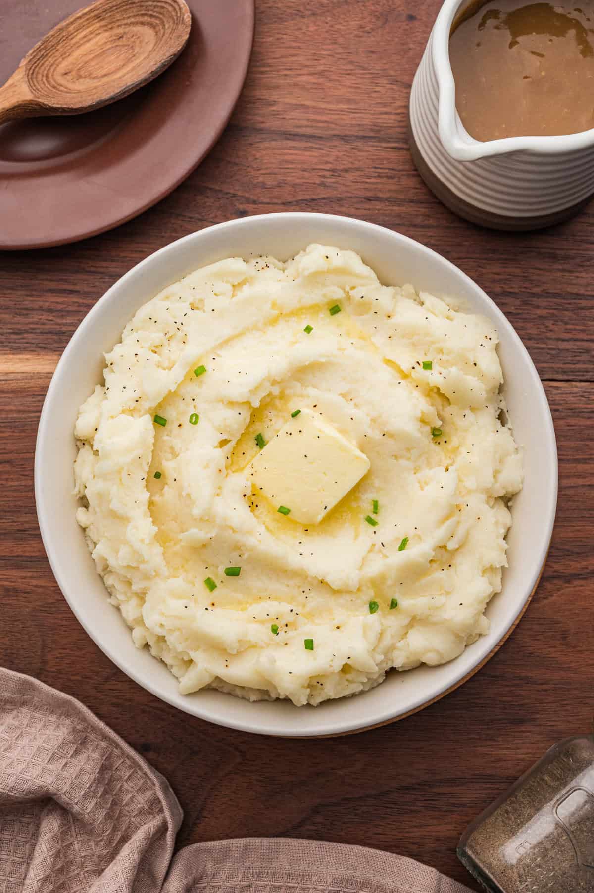 Overhead shot of a bowl of old fashioned mashed potatoes topped with butter, pepper, and chives. The bowl is on a wooden table surrounded by a gravy boat, pepper shaker, wooden spoon, and beige napkins.