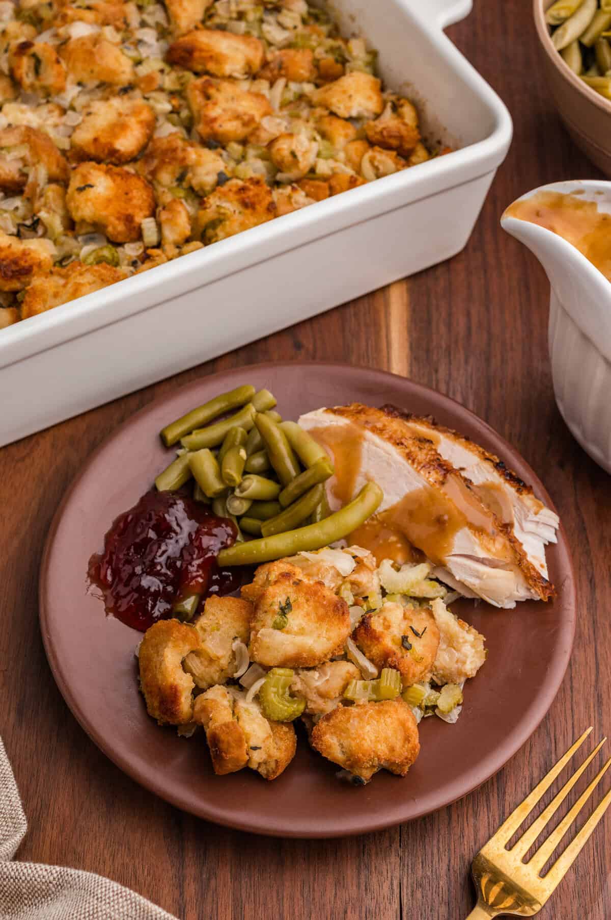 A plate with a serving of golden-brown bread stuffing, slices of turkey breast with gravy, green beans, and cranberry sauce. In the background, a white baking dish of stuffing and a gravy boat are partially visible on a wooden table.