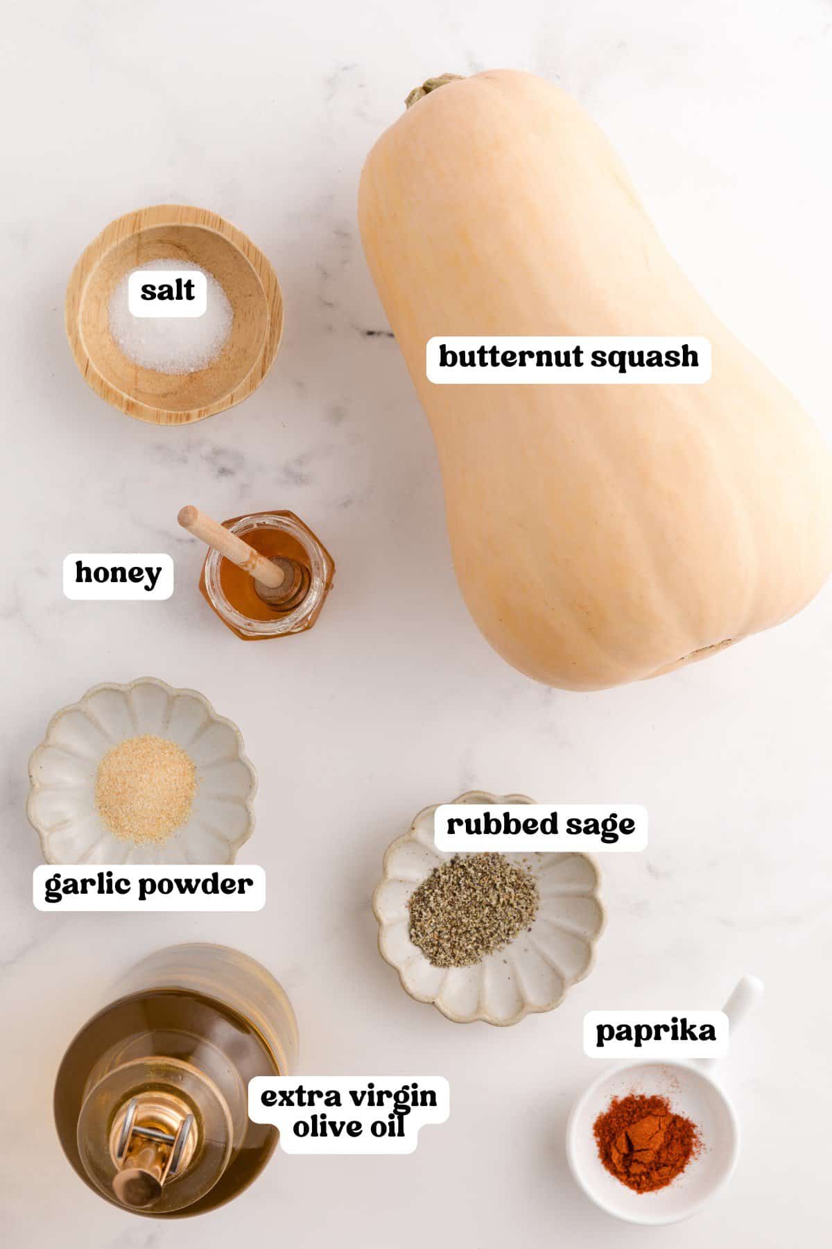 Labeled ingredients for roasted butternut squash, including a whole butternut squash, salt, honey, garlic powder, rubbed sage, paprika, and a bottle of extra virgin olive oil.