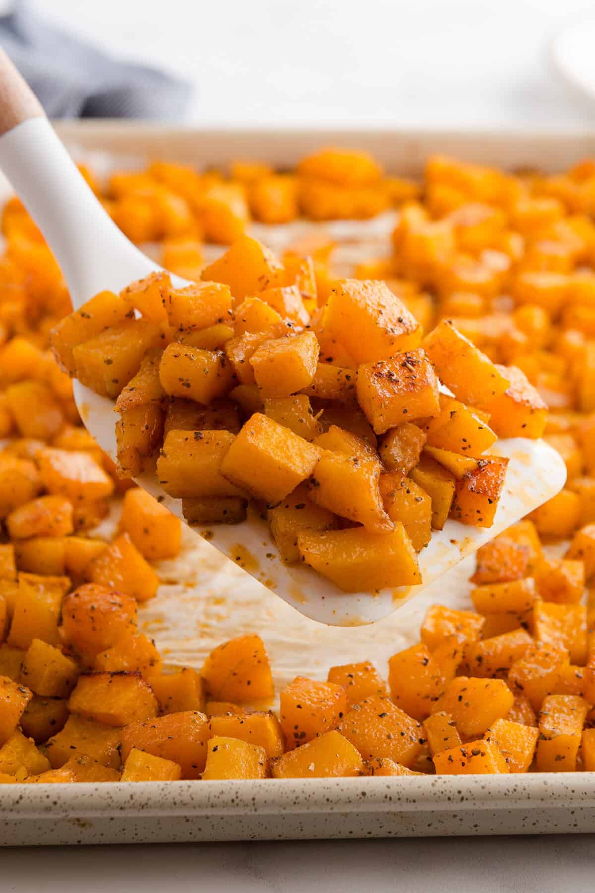 A spatula lifting a portion of roasted butternut squash cubes from a parchment-lined baking sheet. The squash is golden and seasoned, with the rest of the tray visible in the background.
