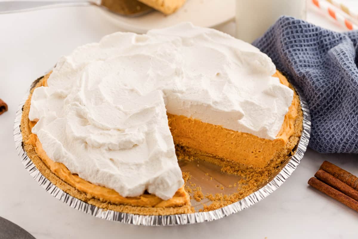 A full no-bake pumpkin pie in a foil pan with a graham cracker crust and a thick layer of whipped topping. One slice has been removed, revealing the creamy pumpkin filling.