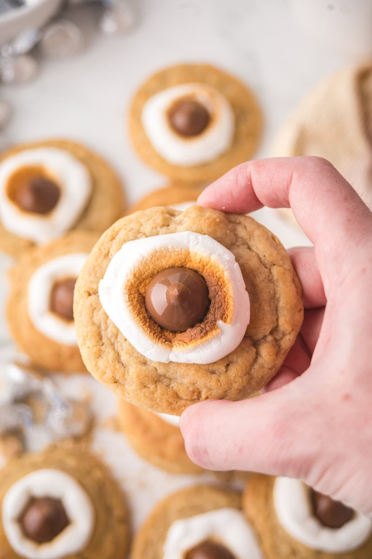 Hand holding a s'mores blossom cookie with toasted marshmallow and chocolate hershey kiss in the center.