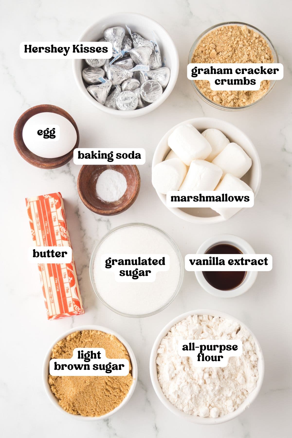 Overhead view of labeled ingredients for s'mores cookies. Items include a bowl of Hershey Kisses, graham cracker crumbs, marshmallows, all-purpose flour, light brown sugar, granulated sugar, butter, vanilla extract, baking soda, and an egg, all arranged on a white surface in separate bowls or containers.