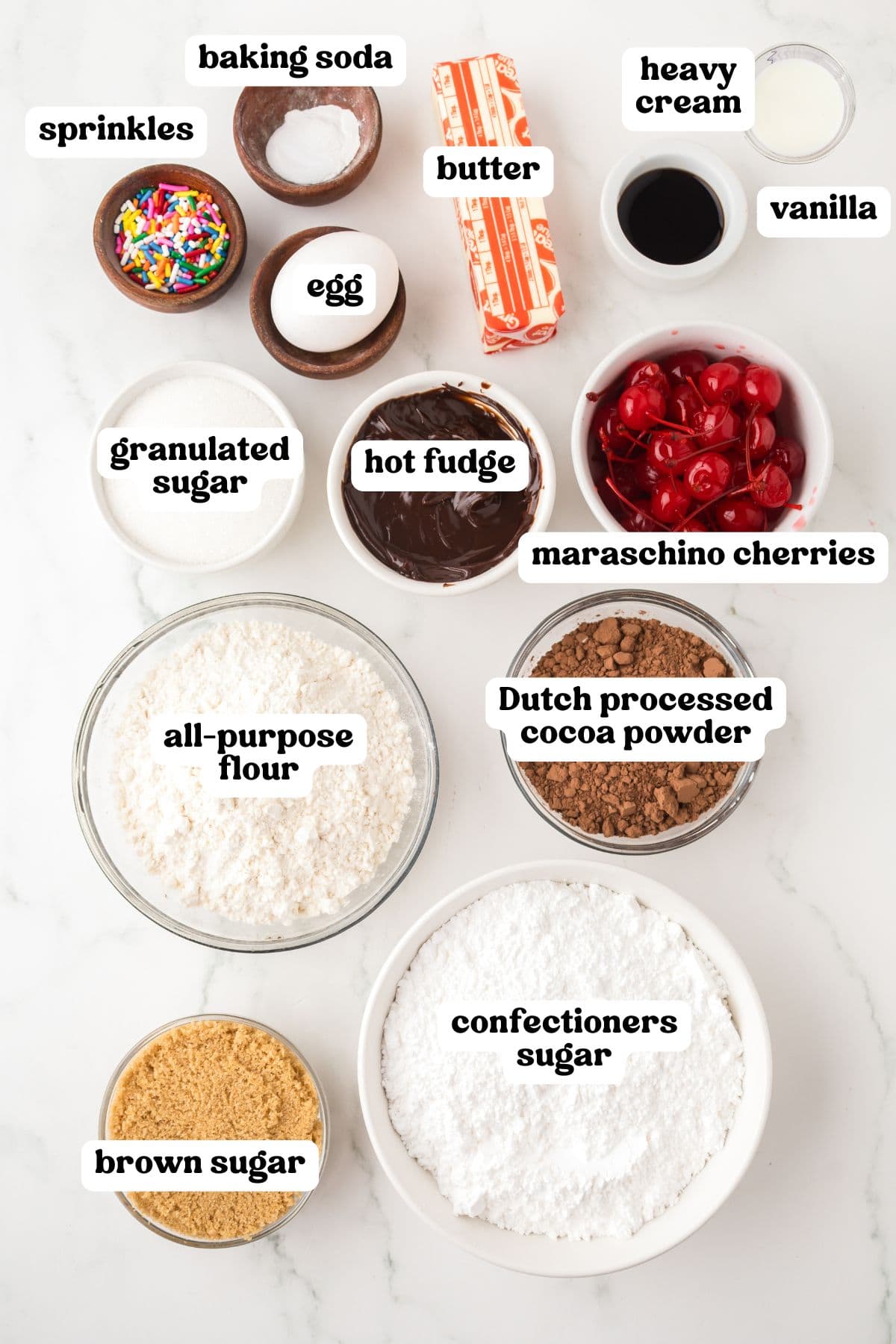 Overhead view of labeled ingredients, including sprinkles, baking soda, butter, heavy cream, vanilla, egg, granulated sugar, hot fudge, maraschino cherries, all-purpose flour, Dutch processed cocoa powder, brown sugar, and confectioners sugar.