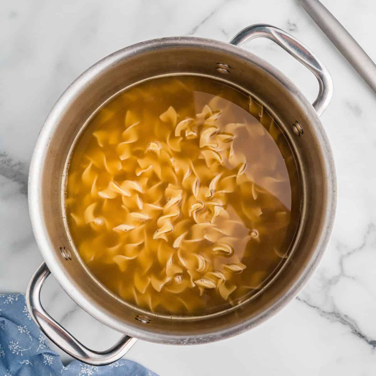 Egg noodles cooking in broth in a stainless steel pot.