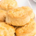Overhead view of multiple golden brown biscuits piled on a white serving platter, with a honey dipper, bowl of honey, and stacked plates visible in the background.