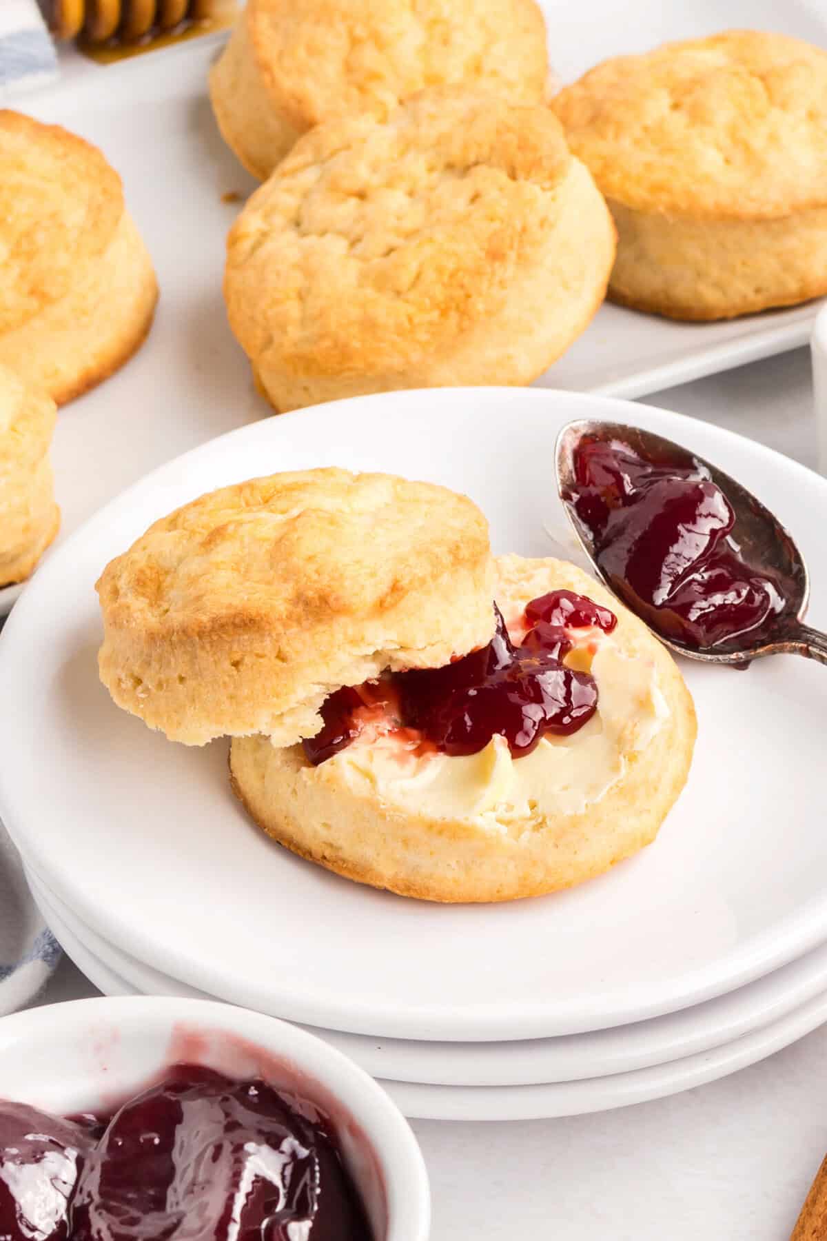 Golden biscuit cut in half and topped with butter and jam, placed on a stack of white plates with more biscuits and jam bowls in the background.